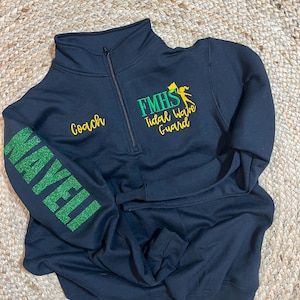May include: A navy blue quarter-zip sweatshirt with the text "Coach" in gold and "NAYELI" in green glitter on the sleeve. The front features "FMHS Tidal Wave Guard" in green and yellow. The sweatshirt is folded on a woven surface.