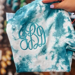 May include: A white and teal tie-dye t-shirt with a green embroidered monogram. The monogram is a stylized "BB" with a small circle in the center.
