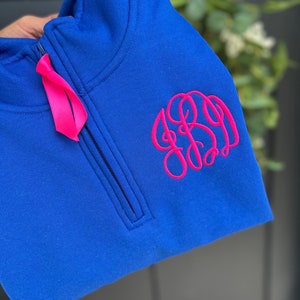 May include: A royal blue quarter-zip sweatshirt with a pink monogram on the right side.