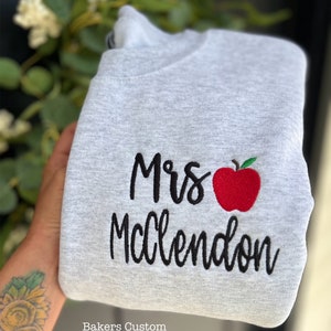 May include: Gray sweatshirt with black embroidery that says "Mrs. McClendon" with a red apple next to the name. The text "Bakers Custom Apparel" is at the bottom of the image.