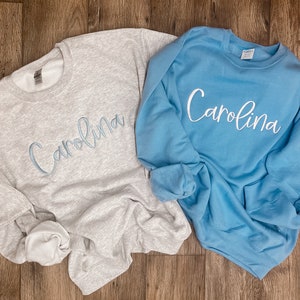 May include: Two sweatshirts, one grey and one light blue, with the name "Carolina" embroidered on each in a light blue cursive font.