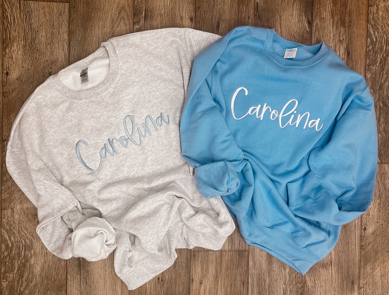 May include: Two sweatshirts, one grey and one light blue, with the name "Carolina" embroidered on each in a light blue cursive font.