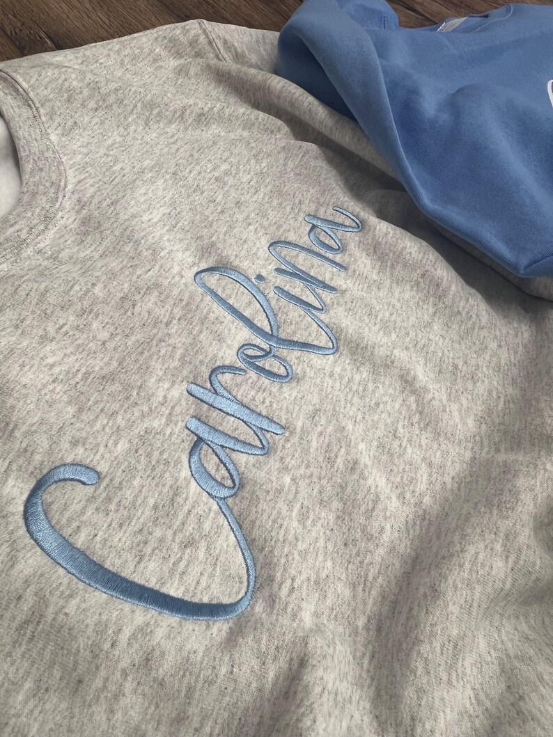May include: A grey sweatshirt with the word "Carolina" embroidered in light blue cursive lettering.