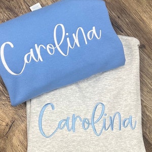May include: Two sweatshirts with the name "Carolina" embroidered on them. The top sweatshirt is light blue with white embroidery. The bottom sweatshirt is grey with light blue embroidery.