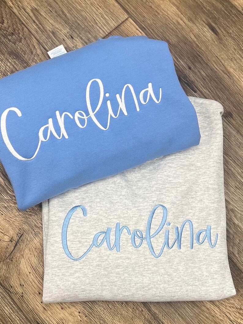 May include: Two sweatshirts with the name "Carolina" embroidered on them. The top sweatshirt is light blue with white embroidery. The bottom sweatshirt is grey with light blue embroidery.