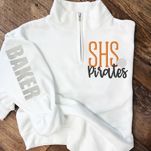 May include: White quarter-zip sweatshirt with the text "SHS Pirates" in orange and black embroidery on the front. The left sleeve has the word "BAKER" in silver glitter.