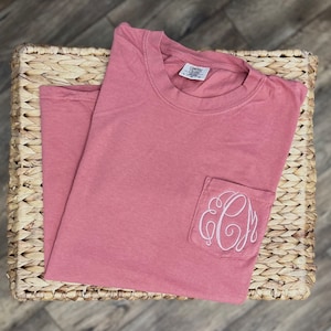 May include: A dusty rose colored t-shirt with a single pocket on the left side. The pocket has a white embroidered monogram with the letters "CJM".