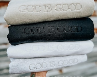 Puede incluir: Una pila de cuatro sudaderas con el texto "GOD IS GOOD" bordado en cada una. Las sudaderas son de diferentes colores: beige, negro, blanco y gris jaspeado. El texto "BAKERS CUSTOM APPAREL" es visible en la parte inferior de la imagen.