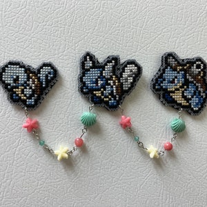 May include: Three handmade cross-stitch brooches featuring a blue and white cartoon character with a light blue, pink, and yellow beaded chain.