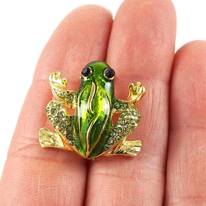 May include: A green and gold frog brooch with rhinestones. The frog is sitting on its hind legs with its front legs raised. The brooch is approximately 2.5 cm tall.