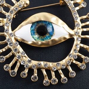 May include: A gold-tone brooch with a large blue eye design surrounded by clear rhinestones. The eye is white with a blue iris and black pupil.