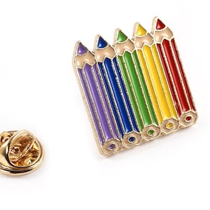 Peut inclure: Une épingle dorée avec un motif de sept crayons de couleur. Les crayons sont disposés en rangée, les extrémités des gommes face au spectateur. Les crayons sont de couleur violette, bleue, verte, jaune, orange, rouge et rose.