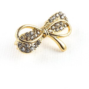 May include: A gold-toned bow brooch with clear rhinestones. The bow is tied in a knot with the ends of the bow extending out to the sides.