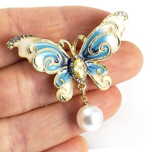 May include: A gold-tone butterfly brooch with blue and white enamel and a dangling pearl. The butterfly has intricate details and a clear rhinestone in the centre.