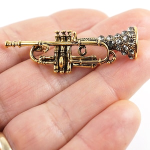 Puede incluir: Un amuleto de trompeta dorado con brillantes transparentes en la campana. El amuleto está en una mano.