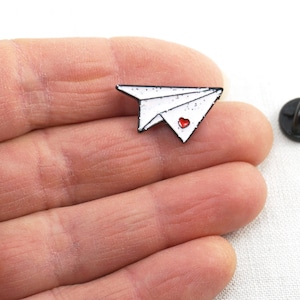 May include: White enamel paper airplane pin with a red heart on the front. The pin is on a hand and the back of the pin is visible.