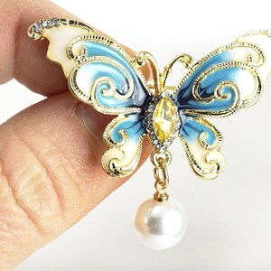May include: A gold-tone butterfly brooch with blue and white enamel and a dangling pearl. The butterfly has intricate details and a clear rhinestone in the centre.