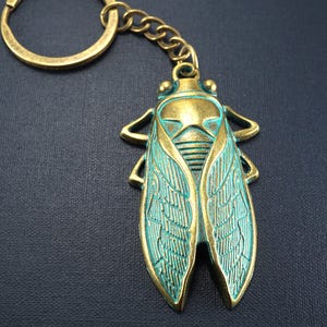 May include: A bronze-colored cicada keychain with a key ring and chain. The cicada has teal-colored wings and a detailed design. The keychain is made of metal and is on a dark blue background.