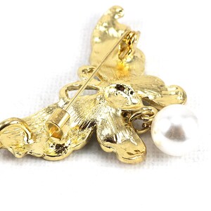 May include: A gold butterfly brooch with a dangling pearl. The butterfly has intricate details and a textured finish.