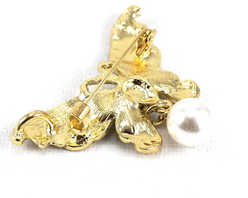 May include: A gold butterfly brooch with a dangling pearl. The butterfly has intricate details and a textured finish.