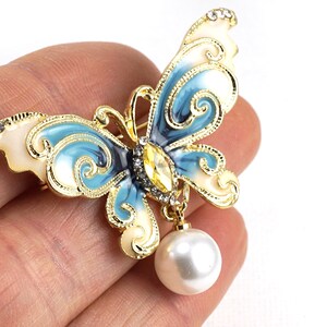 May include: A gold-tone butterfly brooch with blue and white enamel and a dangling pearl. The butterfly has intricate details and a clear rhinestone in the centre.