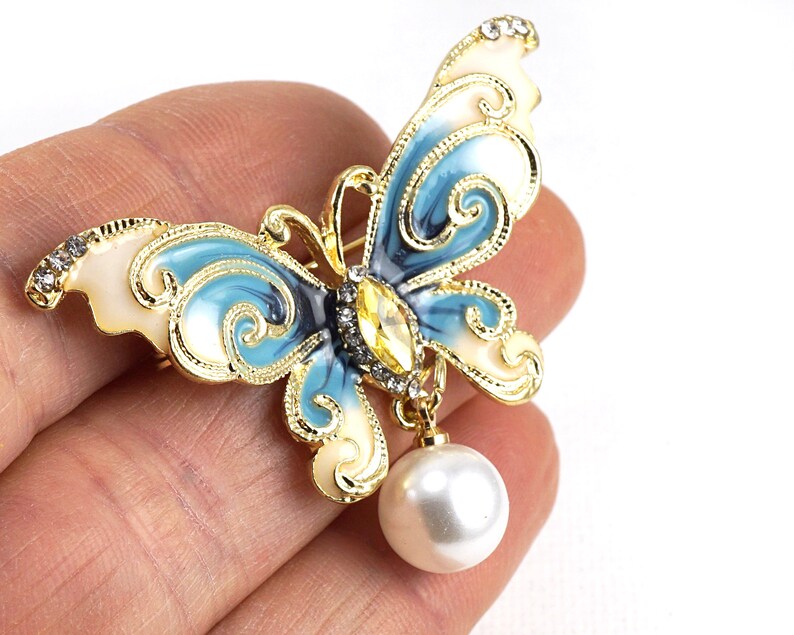 May include: A gold-tone butterfly brooch with blue and white enamel and a dangling pearl. The butterfly has intricate details and a clear rhinestone in the centre.