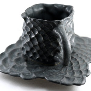 May include: A black ceramic teacup and saucer set with a textured, scaly design. The cup and saucer have a unique, organic shape.