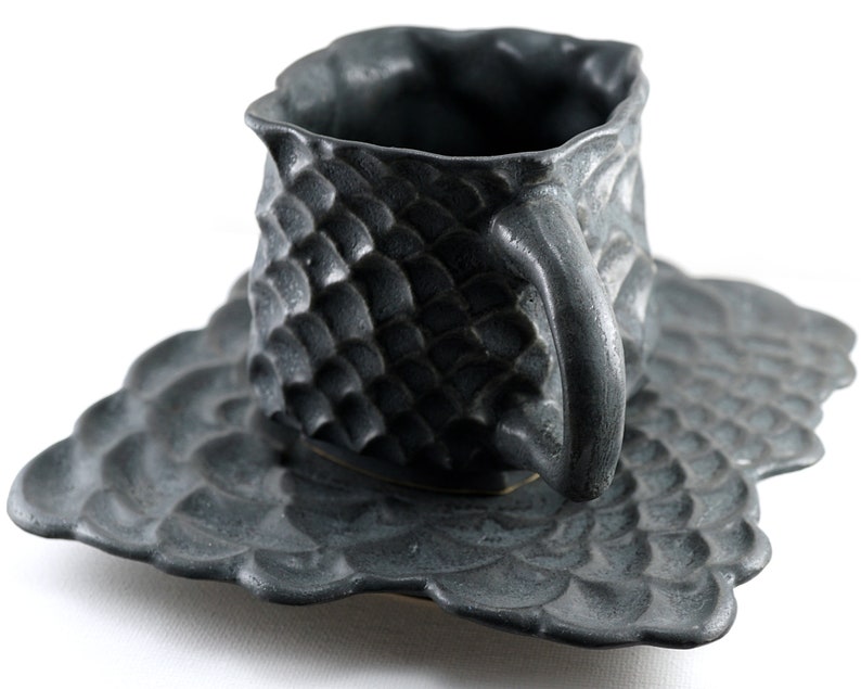 May include: A black ceramic teacup and saucer set with a textured, scaly design. The cup and saucer have a unique, organic shape.