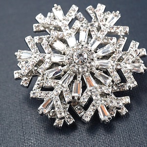 May include: A silver-toned snowflake brooch, encrusted with clear, sparkling crystals. The brooch features a central, round crystal surrounded by rectangular and smaller crystals, creating a dazzling winter-themed accessory.