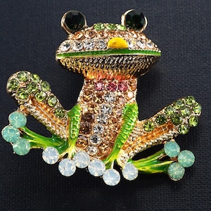 May include: A frog brooch made of gold-tone metal and adorned with rhinestones. The frog has green, clear, and colourful gems, with green and white accents. The eyes are dark green, and the mouth is yellow. The brooch is set against a dark background.