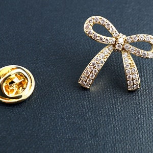 May include: A gold-toned bow-shaped brooch with clear, sparkling stones. The brooch has a pin backing, and is next to a gold-colored pin backing. The brooch is on a dark blue textured surface.