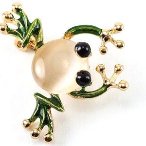 May include: A gold frog brooch with green enamel detailing on the legs and arms. The frog's body is a light beige, round stone, and it has two black eyes. The brooch is a whimsical accessory.