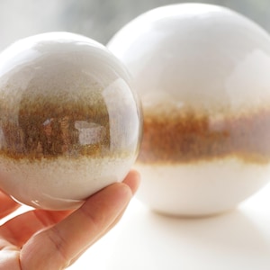 May include: Two ceramic spheres, one held in a hand, the other resting on a surface. Both spheres are white with a brown band around the middle. The smaller sphere is being held in a hand.
