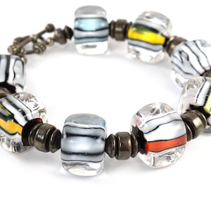 May include: A bracelet with clear glass beads with black and white stripes and accents of yellow, orange, and blue. The beads are separated by silver metal spacers.