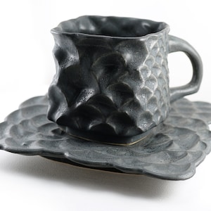 May include: A dark gray ceramic teacup and saucer set with a textured, almost scaly, surface. The cup has a rounded square shape and a handle. The saucer has a scalloped edge.