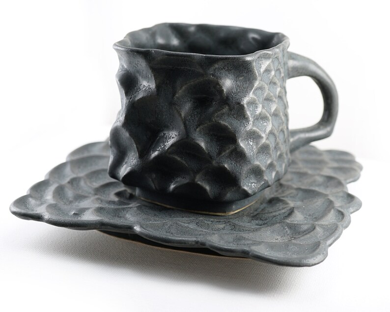 May include: A dark gray ceramic teacup and saucer set with a textured, almost scaly, surface. The cup has a rounded square shape and a handle. The saucer has a scalloped edge.