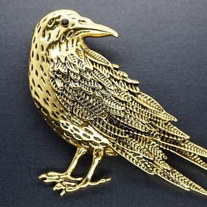 Op de afbeelding: Een goudkleurige vogelbroche met ingewikkelde details. De broche toont een vogel met licht gespreide vleugels, en de veren zijn minutieus weergegeven. De vogel staat op twee poten, en het algehele ontwerp is sierlijk en gedetailleerd.