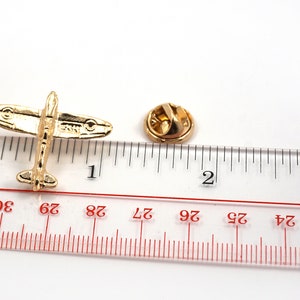 Puede incluir: Pin de metal dorado de un avi&oacute;n de combate de la Segunda Guerra Mundial, que mide aproximadamente 2,5 cm de largo, sobre un fondo blanco junto a una regla.