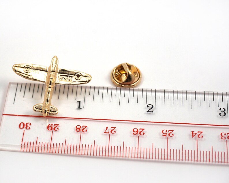 Puede incluir: Pin de metal dorado de un avi&oacute;n de combate de la Segunda Guerra Mundial, que mide aproximadamente 2,5 cm de largo, sobre un fondo blanco junto a una regla.