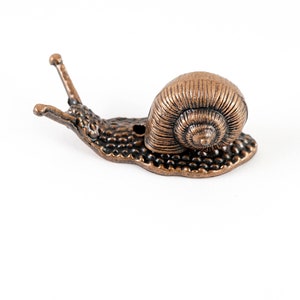 Può includere: Statuetta a forma di lumaca color bronzo con guscio dettagliato e corpo strutturato. Le antenne della lumaca sono estese, su sfondo bianco. Questo oggetto decorativo può essere utilizzato come fermacarte o come elemento d'arredo unico.