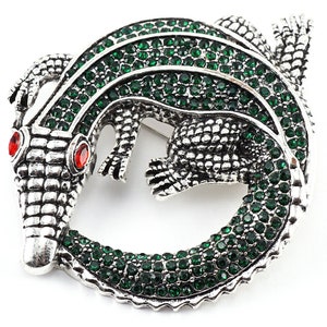 May include: A silver alligator brooch with green rhinestones. The alligator is curled into a circle with its tail in its mouth.