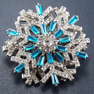May include: A silver-toned snowflake brooch with clear and turquoise-colored crystals. The brooch features a central cluster of clear crystals surrounded by turquoise baguette-cut crystals, with additional clear crystals forming the snowflake's arms.