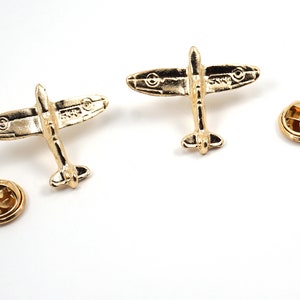 Puede incluir: Dos pines de solapa de metal de color dorado con forma de avi&oacute;n Spitfire. Los pines tienen un dise&ntilde;o detallado y est&aacute;n unidos a una parte posterior circular.