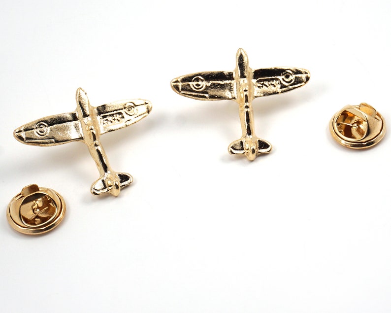 Puede incluir: Dos pines de solapa de metal de color dorado con forma de avi&oacute;n Spitfire. Los pines tienen un dise&ntilde;o detallado y est&aacute;n unidos a una parte posterior circular.