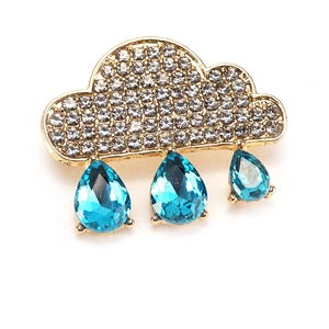 May include: A gold-toned cloud-shaped brooch with a sparkling, crystal-covered top and three dangling, teardrop-shaped, blue gemstones. The brooch is set against a white background, highlighting the contrast of colors and textures.