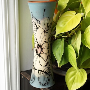 May include: A tall, slender ceramic vase with a light blue glaze and a white and black floral design. The vase has a slightly flared opening and a rounded bottom.