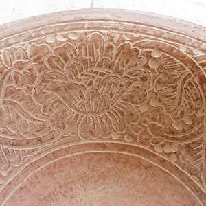 May include: A terracotta bowl with a floral design carved into the surface. The bowl is a light brown color and has a smooth, rounded shape.