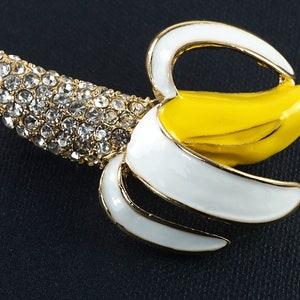 May include: A gold-tone banana brooch with white enamel and yellow enamel. The banana is covered in clear rhinestones.