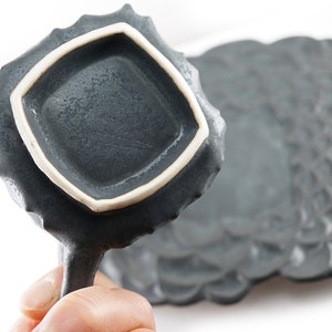 May include: A dark gray ceramic dish with a white rim and a textured surface. The dish has a unique, organic shape and is perfect for serving small snacks or appetizers.