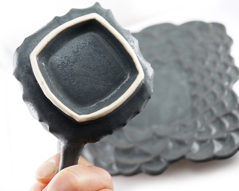 May include: A dark gray ceramic dish with a white rim and a textured surface. The dish has a unique, organic shape and is perfect for serving small snacks or appetizers.